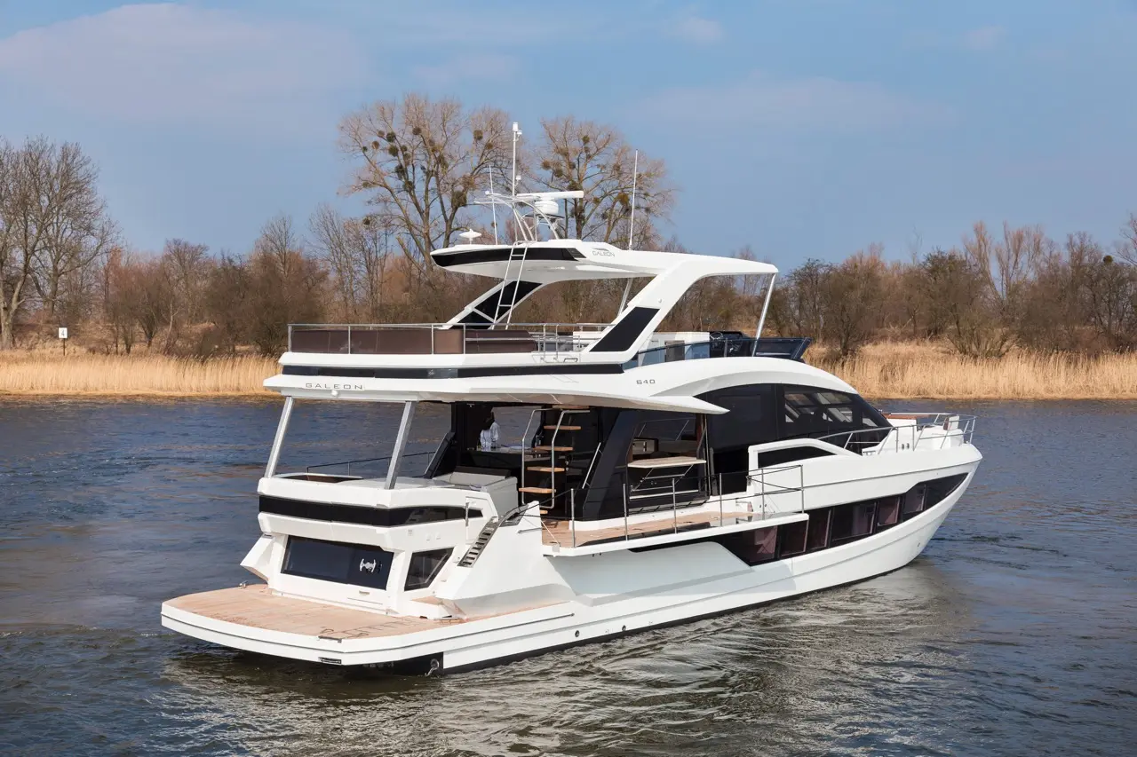 A sleek Galeon 640 yacht cruising on calm waters, surrounded by trees and reeds under a clear blue sky.