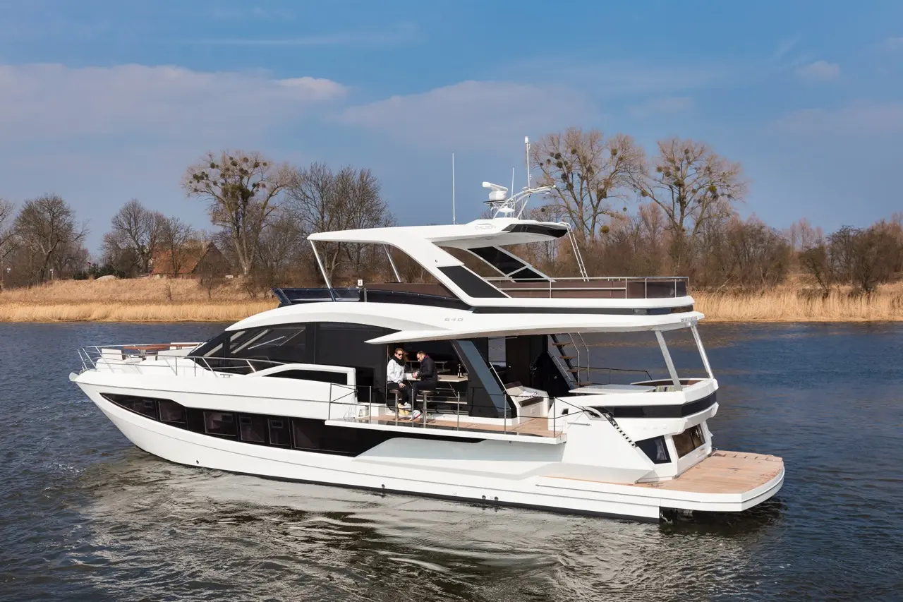 Luxury yacht cruising on a calm waterway, featuring two people enjoying drinks on the deck with trees in the background.