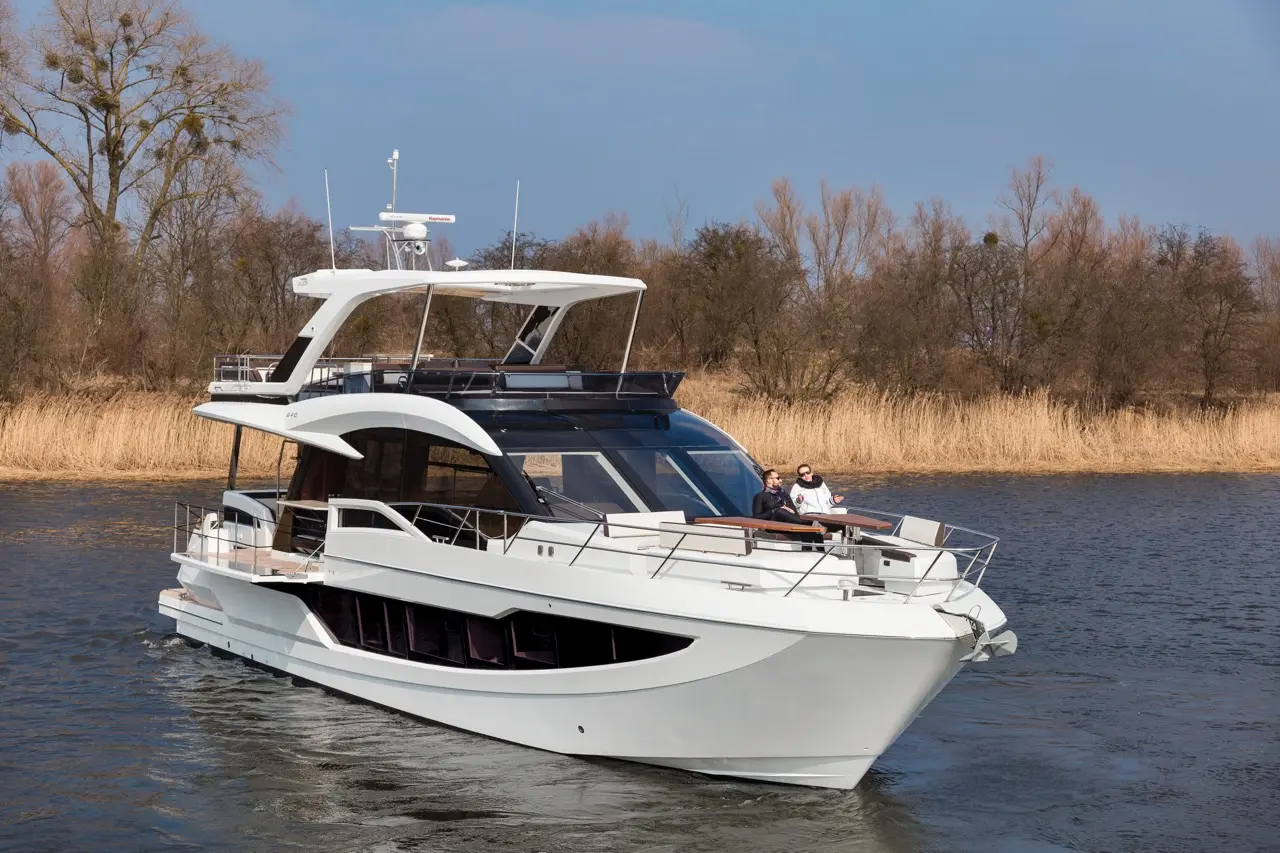 A modern white yacht cruising on a river, with two people relaxing on the deck and trees in the background.