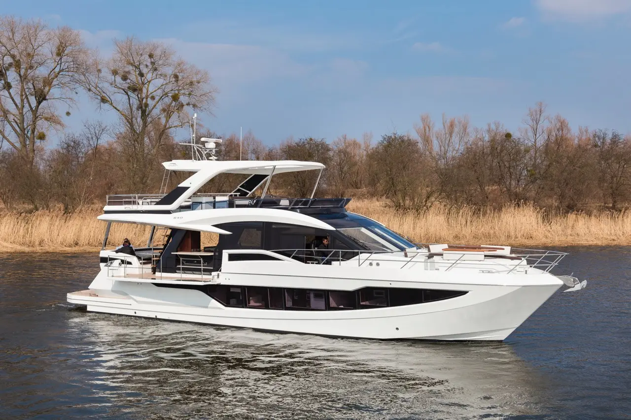 A luxury yacht navigates a calm river, surrounded by trees and reeds under a clear blue sky.