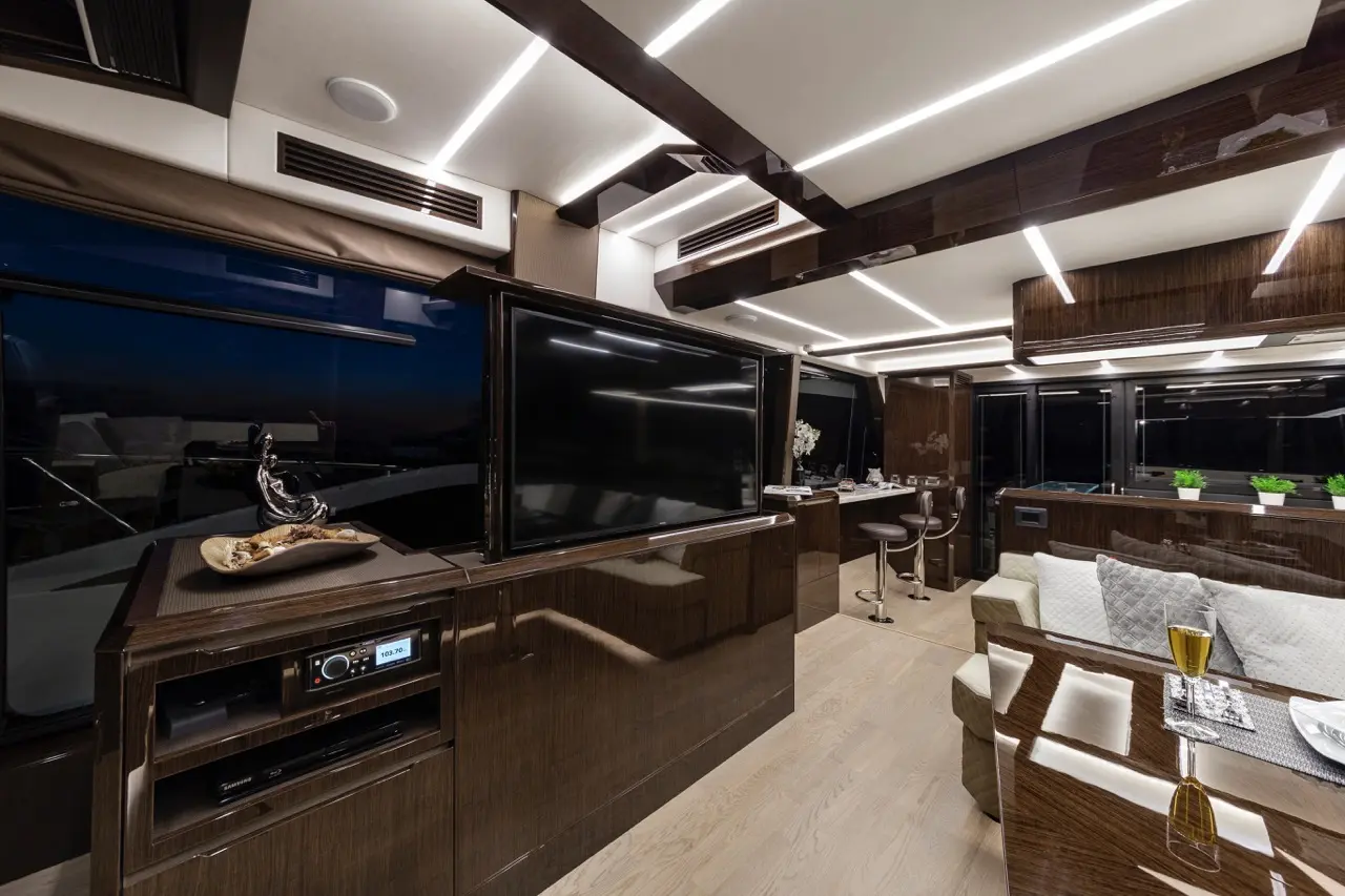 Modern luxury yacht interior with sleek wooden finishes, large TV, and elegant seating, featuring decorative plants and a drink.