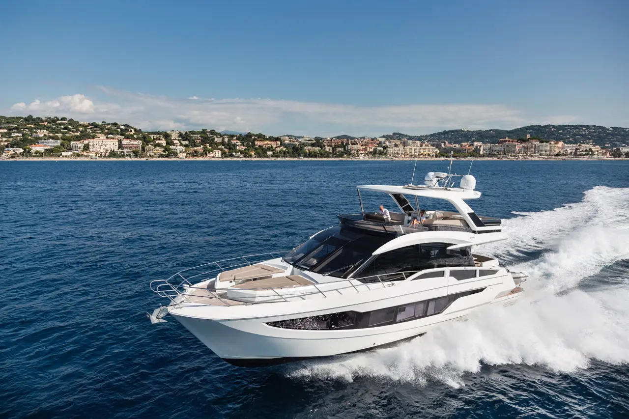 A luxury motor yacht navigates blue waters near a coastal town, showcasing sleek design and a sunny sky.