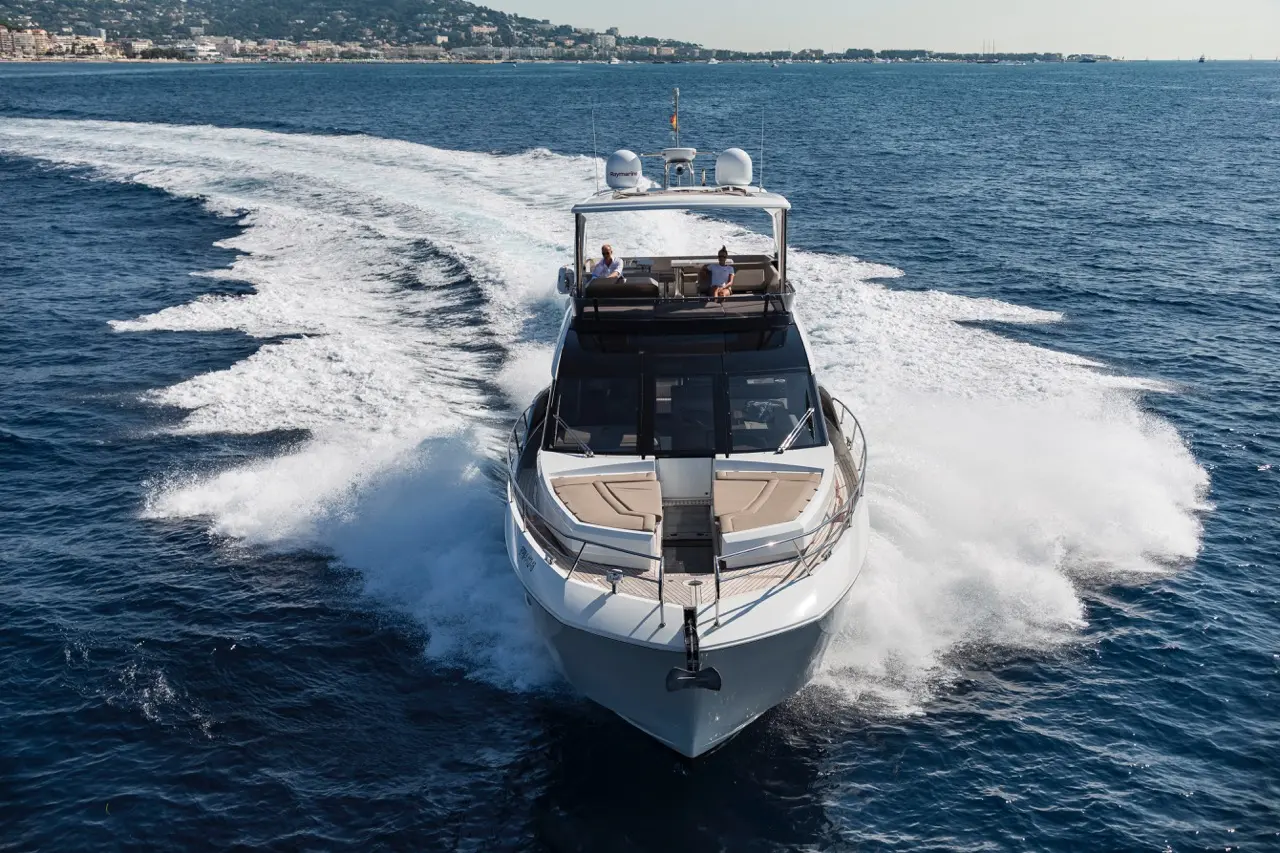 A luxury motorboat cruising on blue waters, leaving a wake, with two people relaxing on deck under sunlight.