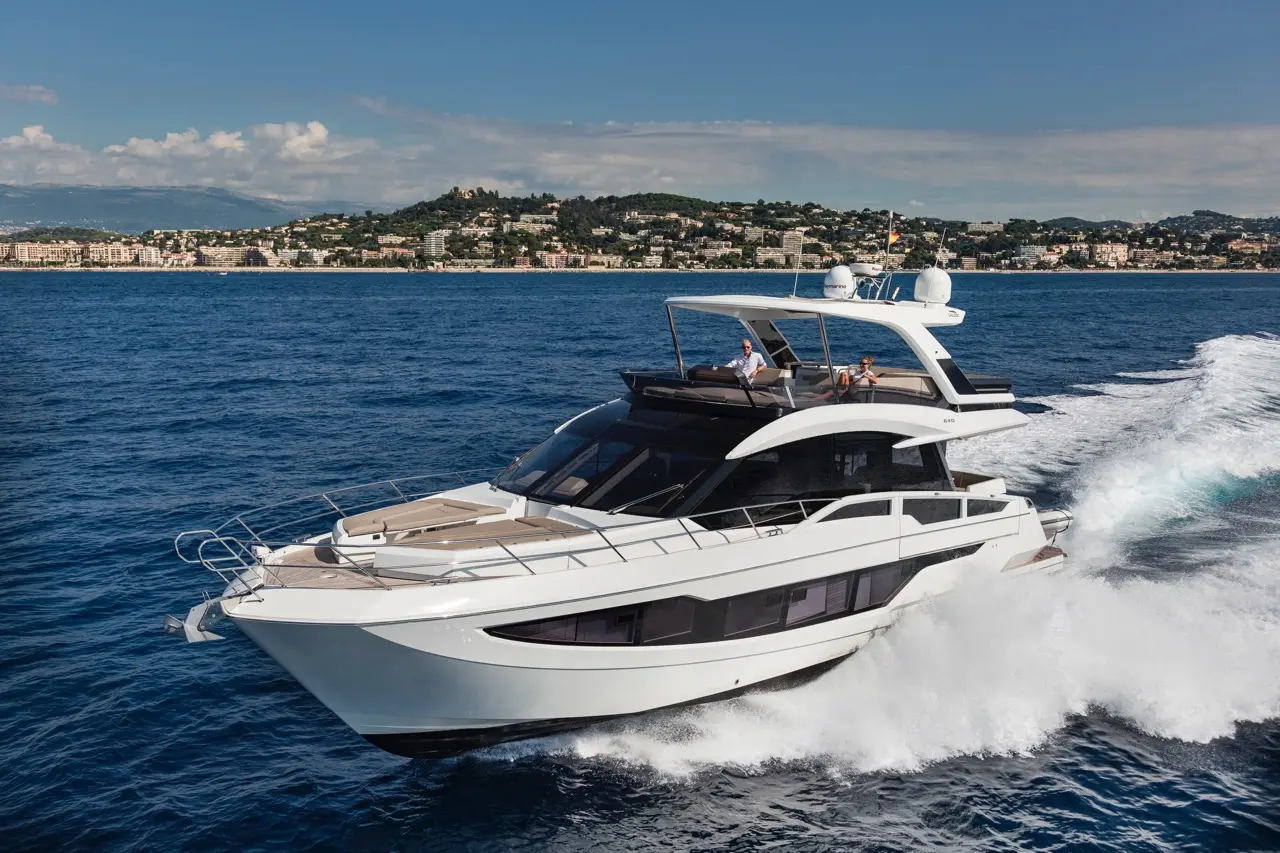 Luxury yacht cruising through blue waters, with two people on deck and a coastal landscape in the background.