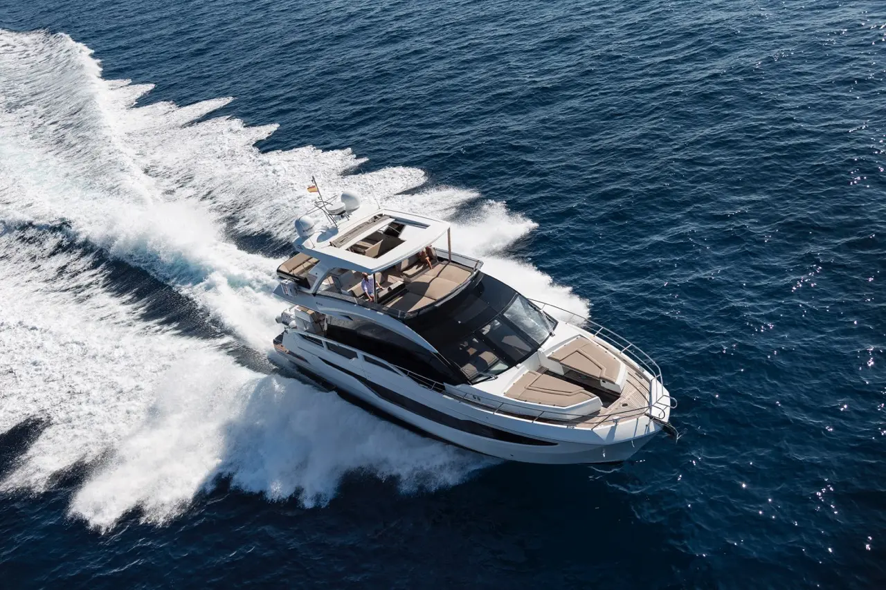 Luxury motor yacht cruising on blue waters, leaving a trail of foam behind. Sunlit deck with seating visible.