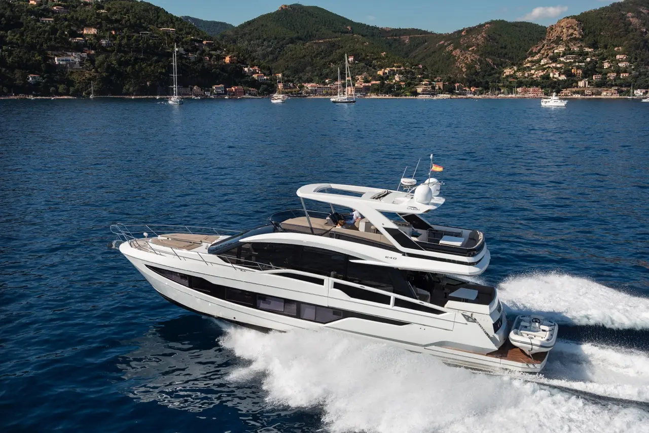 A luxury yacht cruising through clear blue waters, with a mountainous coastline and other boats visible in the background.