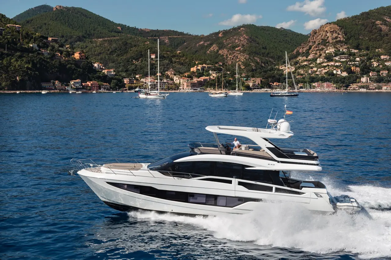 A sleek luxury yacht navigates blue waters, with lush green hills and coastal towns visible in the background.