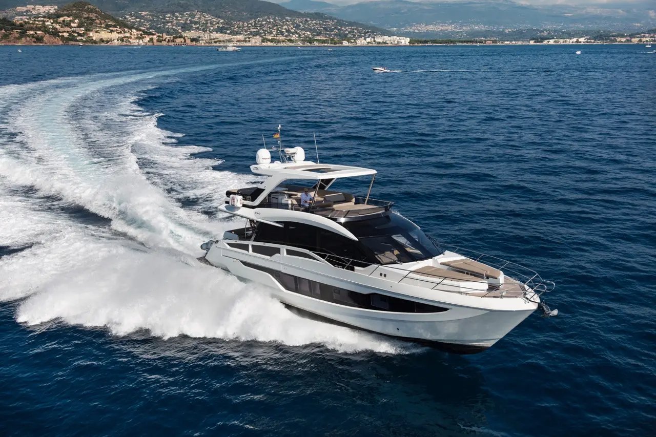 Luxury yacht cruising through blue waters, leaving a white wake, with a scenic coastline and mountains in the background.