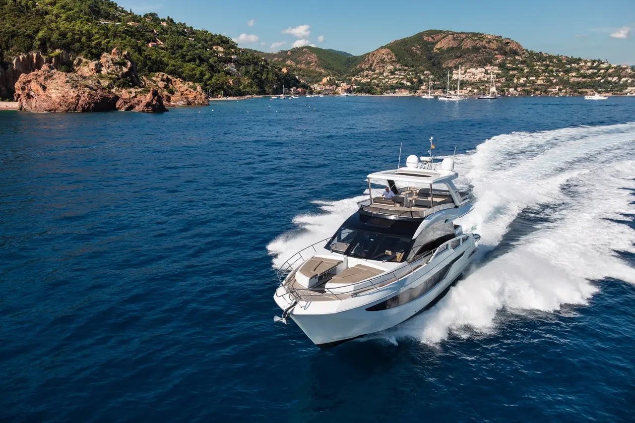 A luxury yacht glides through clear blue waters with scenic hills and a coastline in the background, creating a tranquil scene.