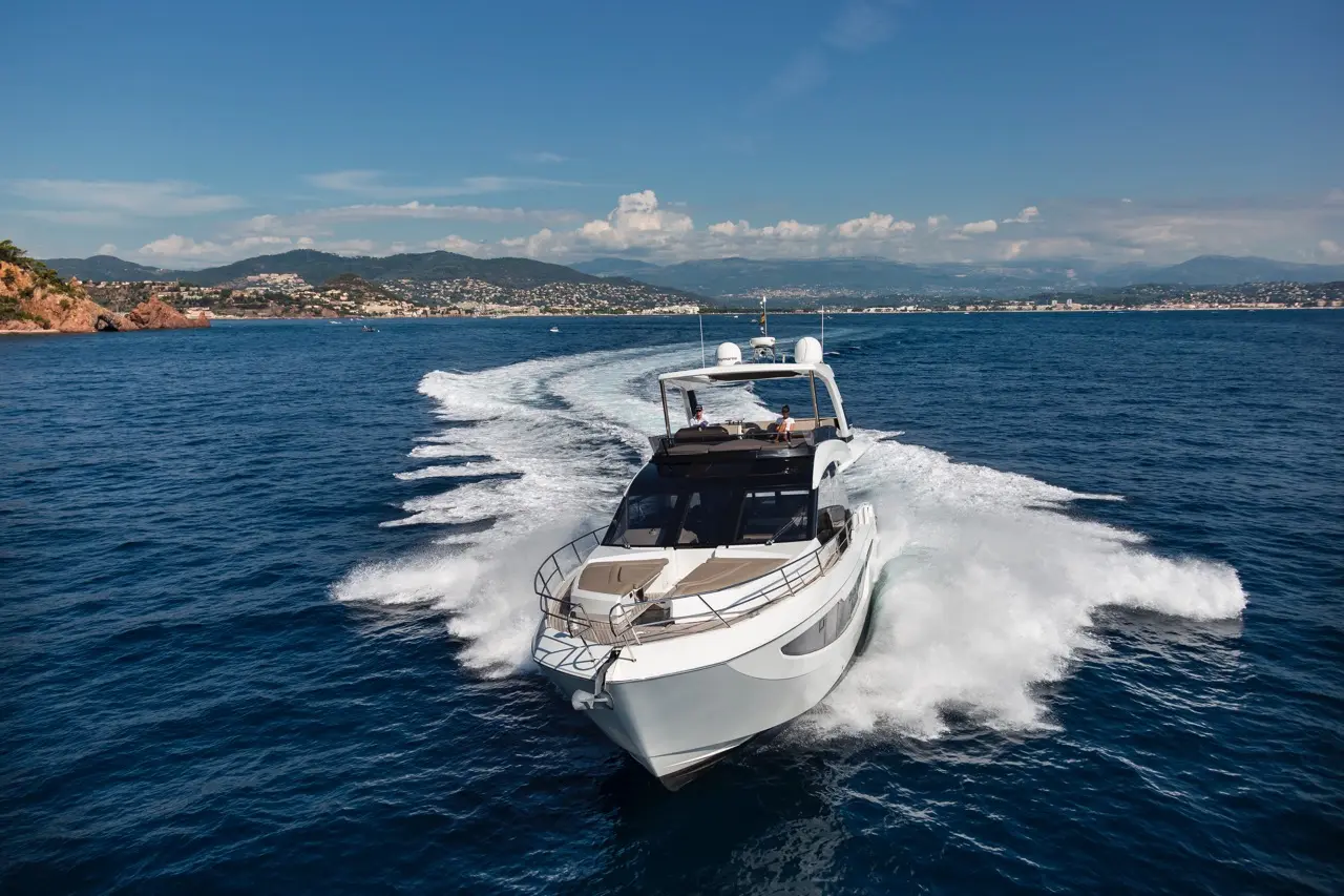 Luxury motor yacht cruising through blue waters, leaving a wake, with scenic coastline and mountains in the background.