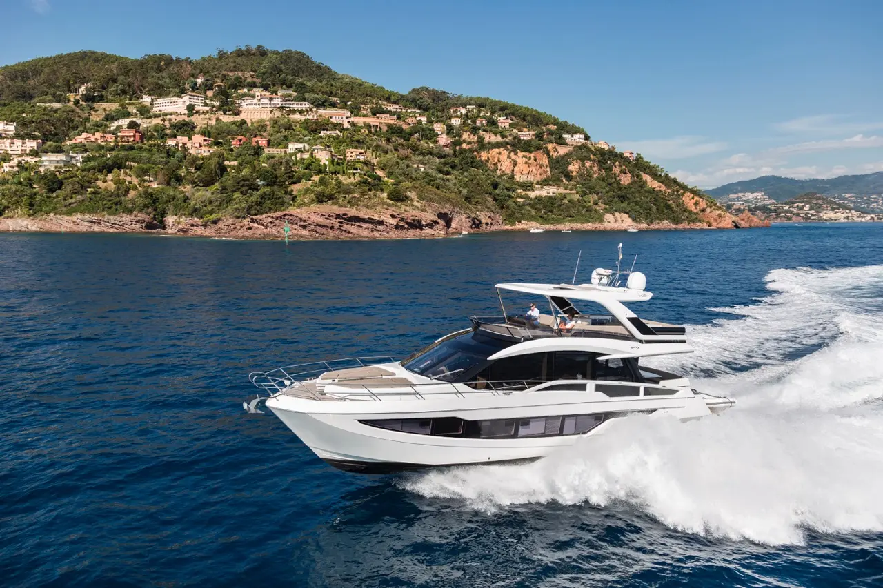 A sleek white yacht navigates blue waters near a lush green hillside with coastal homes, showcasing an upscale marine lifestyle.