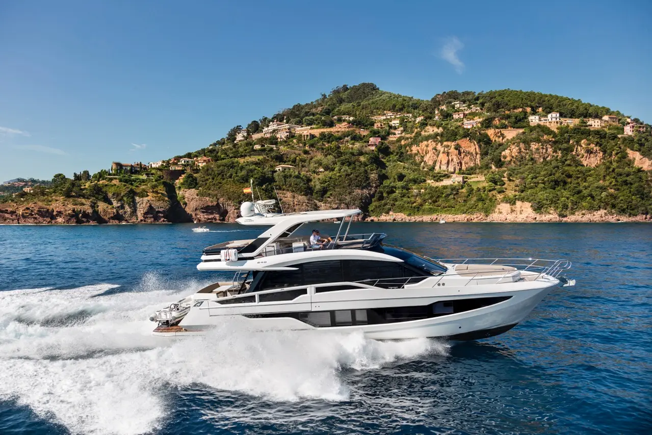 Luxury yacht cutting through clear blue waters, with a scenic coastal backdrop of hills and modern homes.