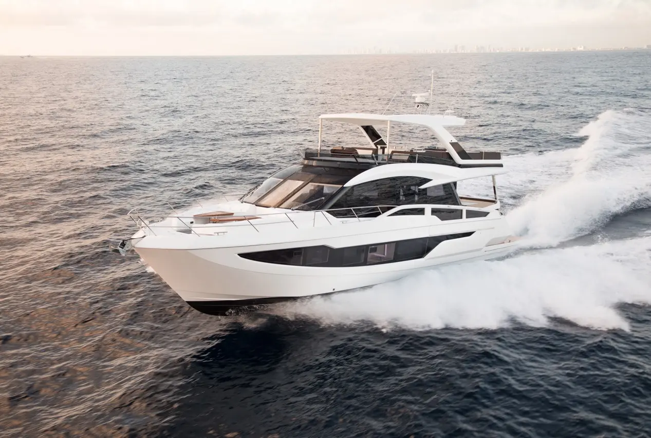A sleek white yacht cruises through calm waters, creating white frothy waves against a backdrop of a hazy horizon.