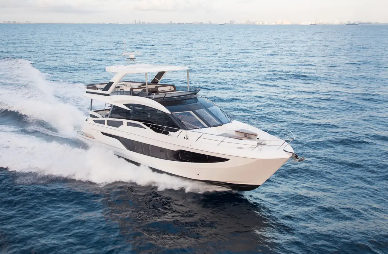 A sleek luxury yacht navigates through blue waters, leaving a wake against a backdrop of distant city skyline.