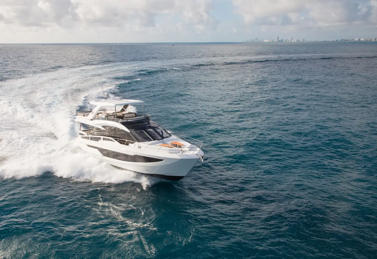 A luxury yacht speeds through clear blue waters, creating waves, with a distant city skyline visible on the horizon.