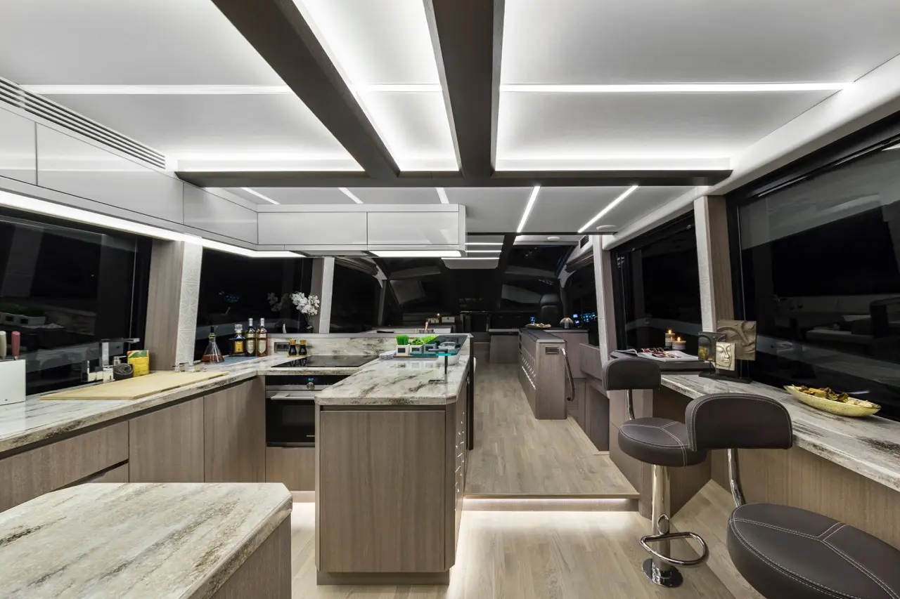 Sleek modern yacht kitchen with wooden cabinetry, marble countertops, bar stools, and ambient lighting, viewed at night.