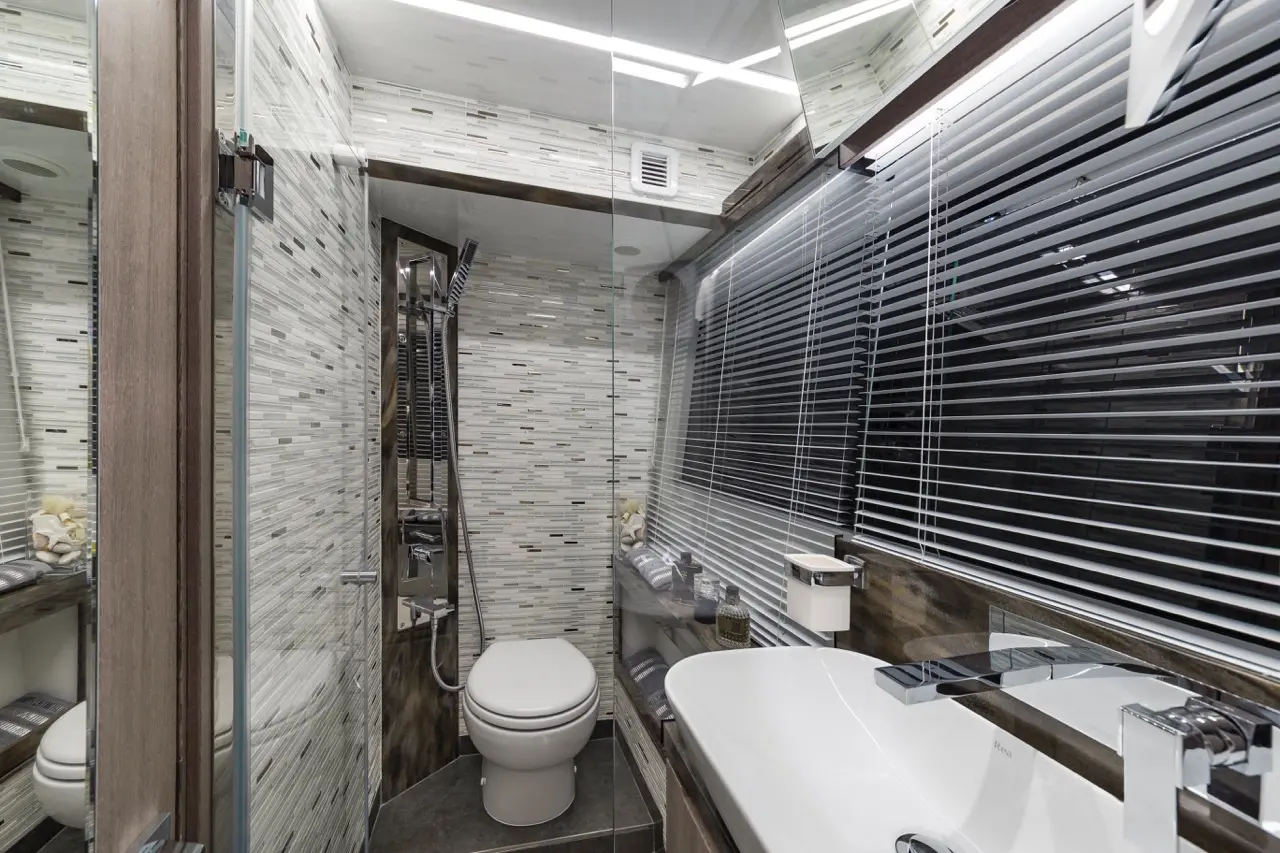 Modern bathroom with glass shower, toilet, large sink, and stylish grey mosaic tiles under bright overhead lighting.