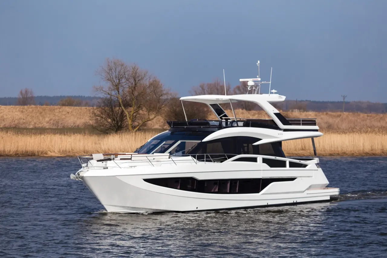 A sleek white yacht cruises on calm waters, with trees and reeds lining the scenic shoreline under a clear blue sky.