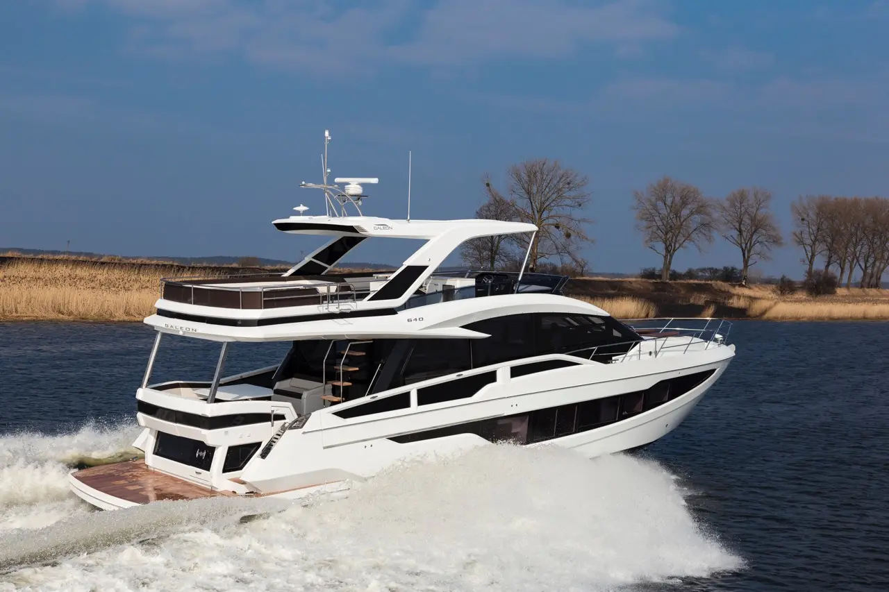 A sleek white yacht, named Galleon 640, glides through water, creating waves with a backdrop of trees and a clear blue sky.