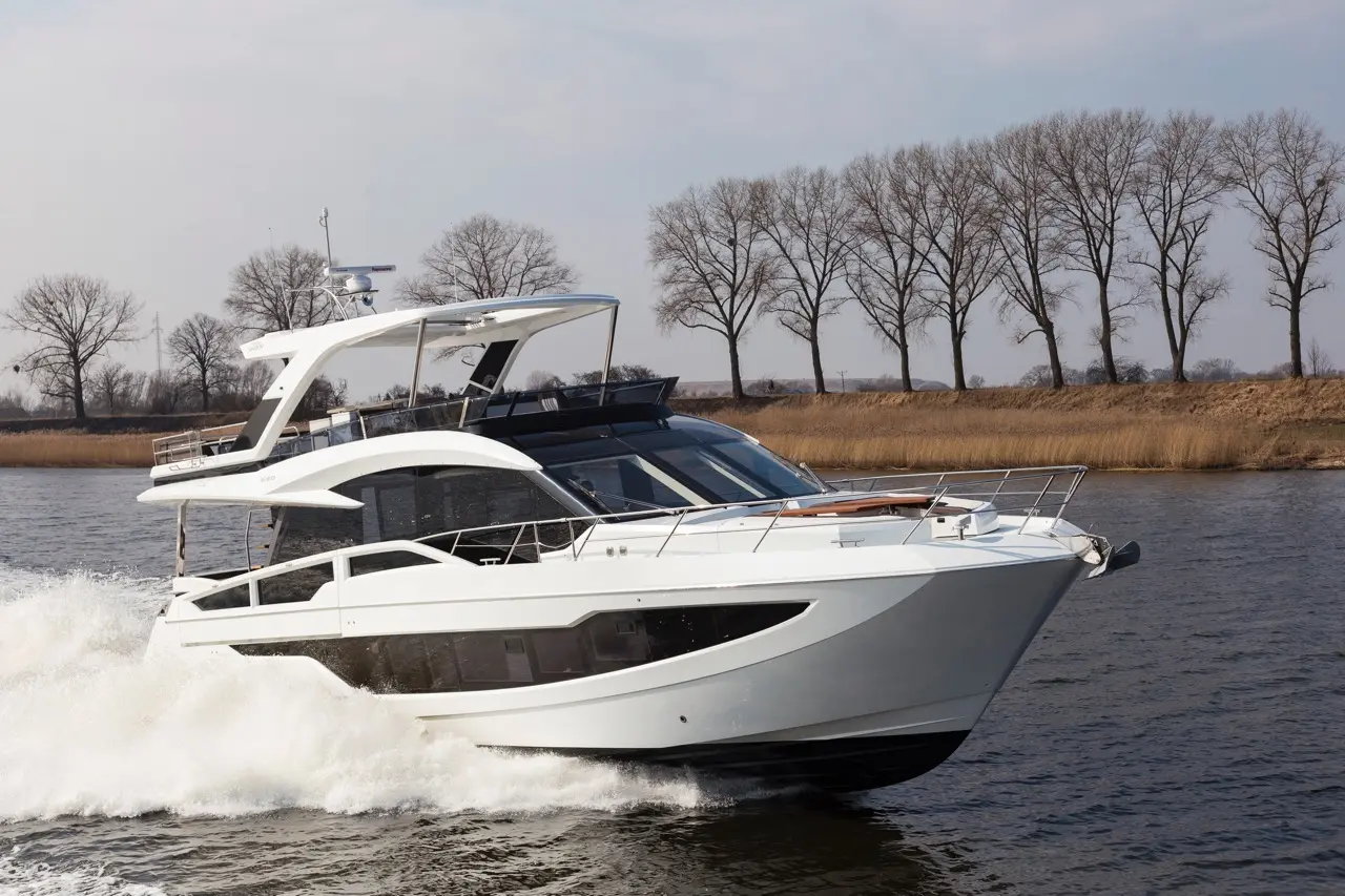 White luxury motor yacht cruising on a river, with splashes of water and trees lining the bank in the background.