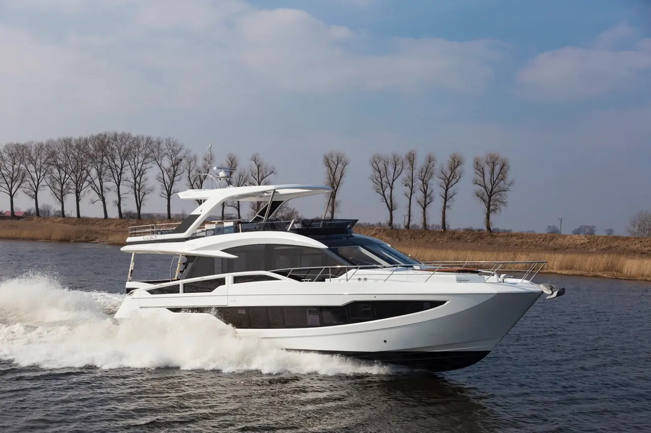 A sleek white yacht navigates through calm waters, surrounded by bare trees and a clear blue sky.