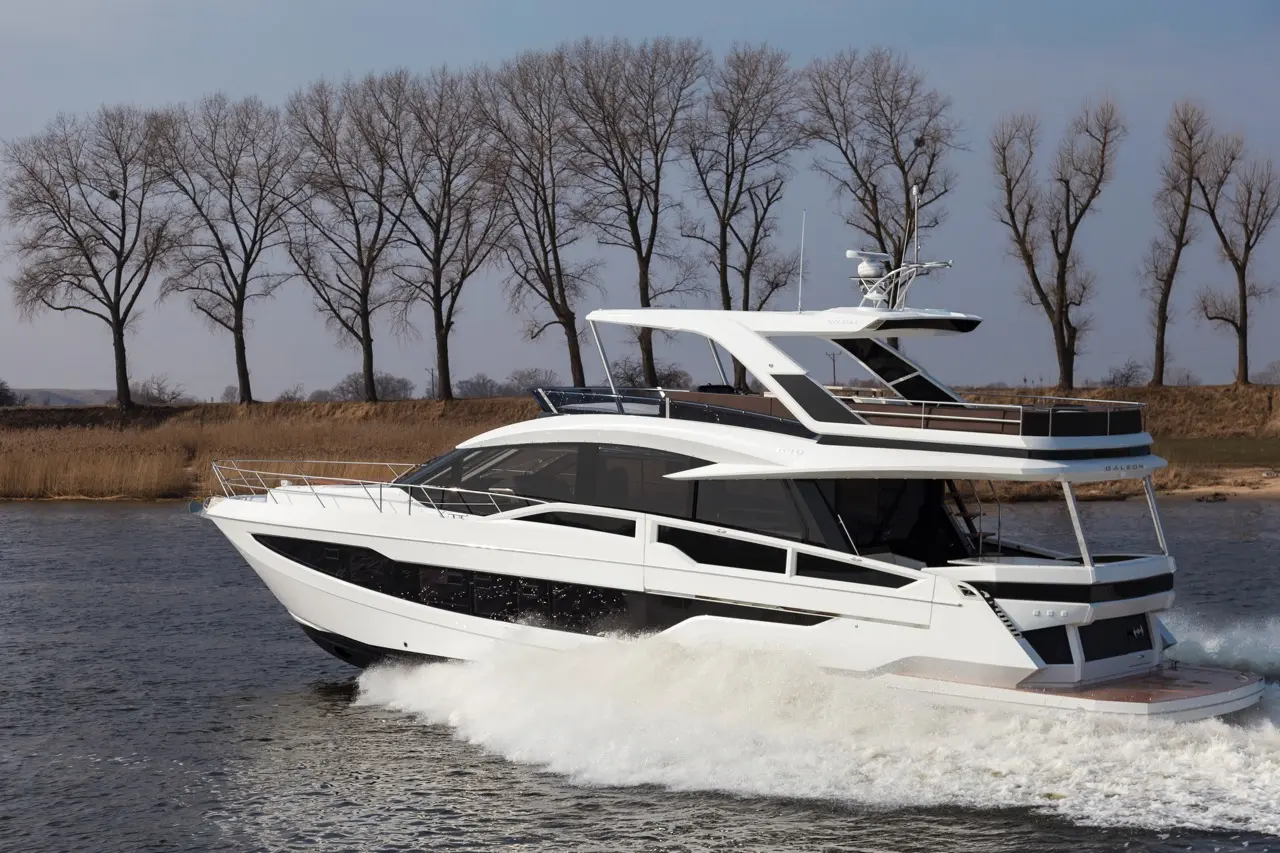 A sleek white yacht glides over water, creating waves, with bare trees lining the shore in a clear sky.