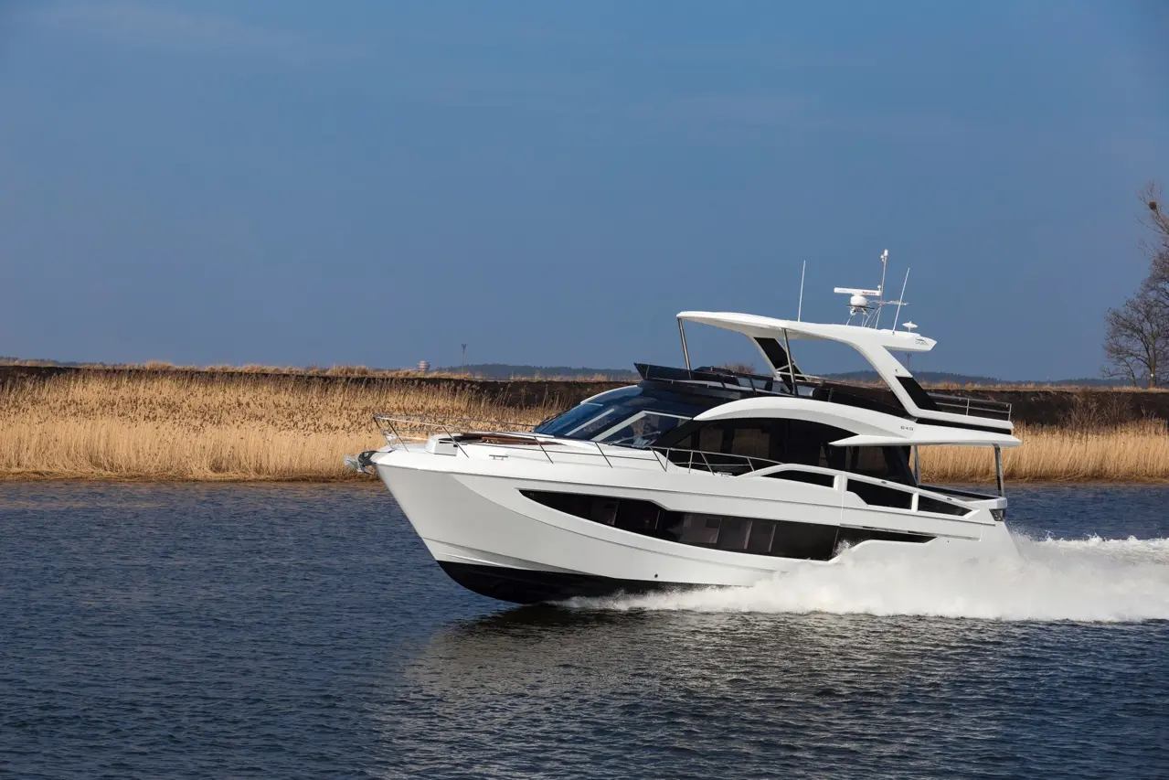 A sleek white motor yacht glides over calm waters, with reeds lining the shore under a clear blue sky.