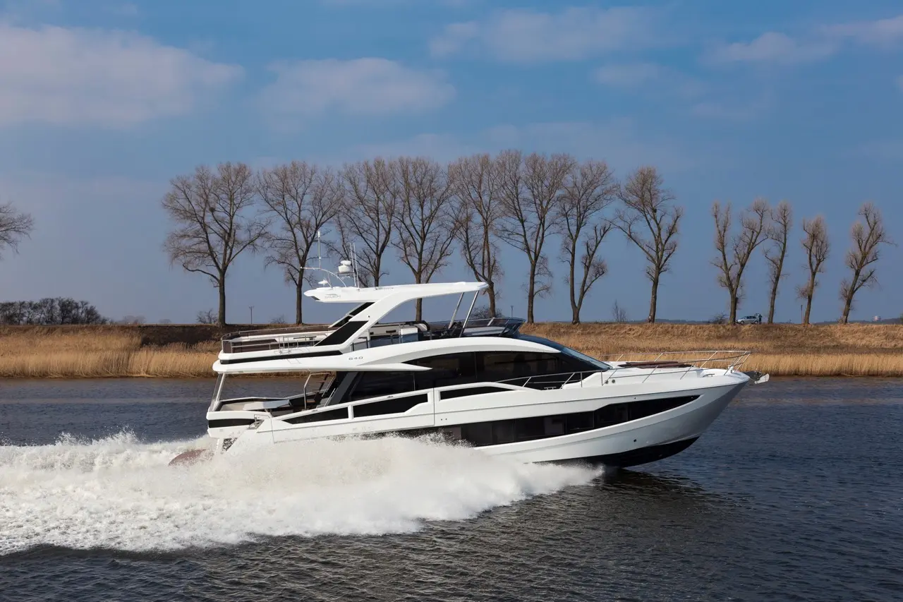 A sleek white yacht speeds across the water, with bare trees lining the shore under a clear blue sky.