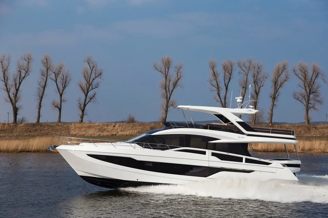 A sleek white yacht navigates through calm waters, with barren trees and a sandy shore in the background under a blue sky.