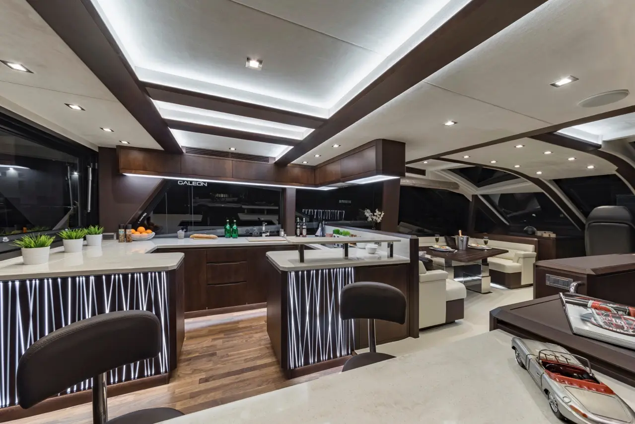 Modern yacht interior featuring a stylish kitchen, wooden counters, bar stools, and a lounge area with elegant decor.