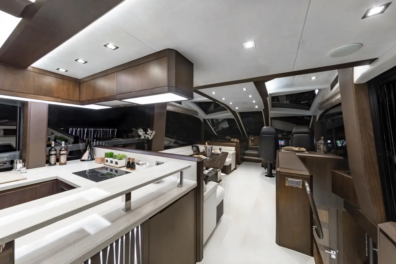 Modern yacht interior featuring a sleek kitchen, lounge area, and helm with panoramic views, illuminated by recessed lighting.