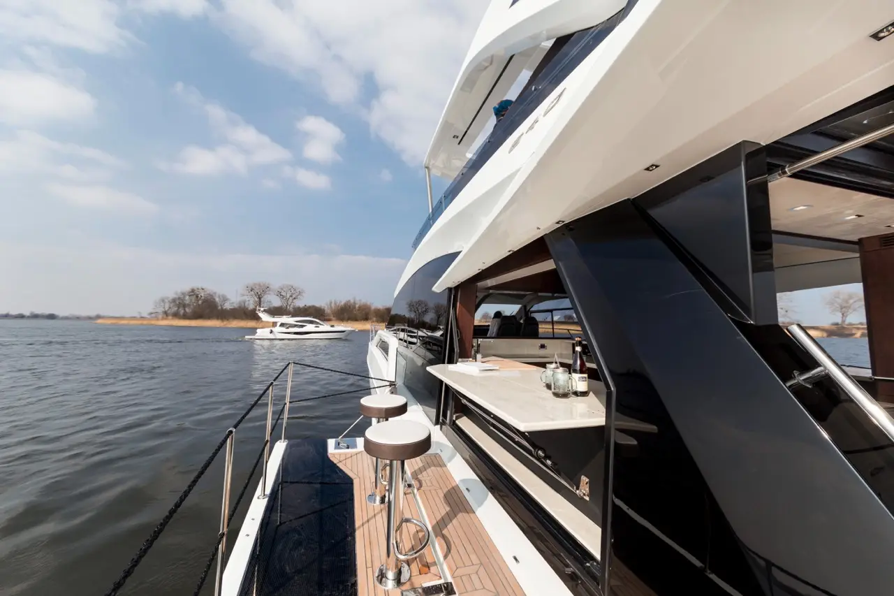 Luxury yacht side view with a bar area, two stools, and scenic waterfront landscape under a blue sky.