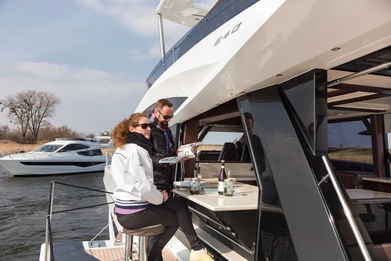 A man and woman enjoy drinks on a luxury yacht, looking at a magazine with water and other boats in the background.