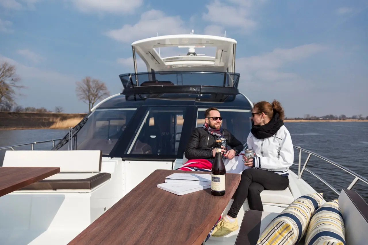 A couple enjoys drinks on a luxury boat, surrounded by water and trees, with a bottle of champagne on the table.