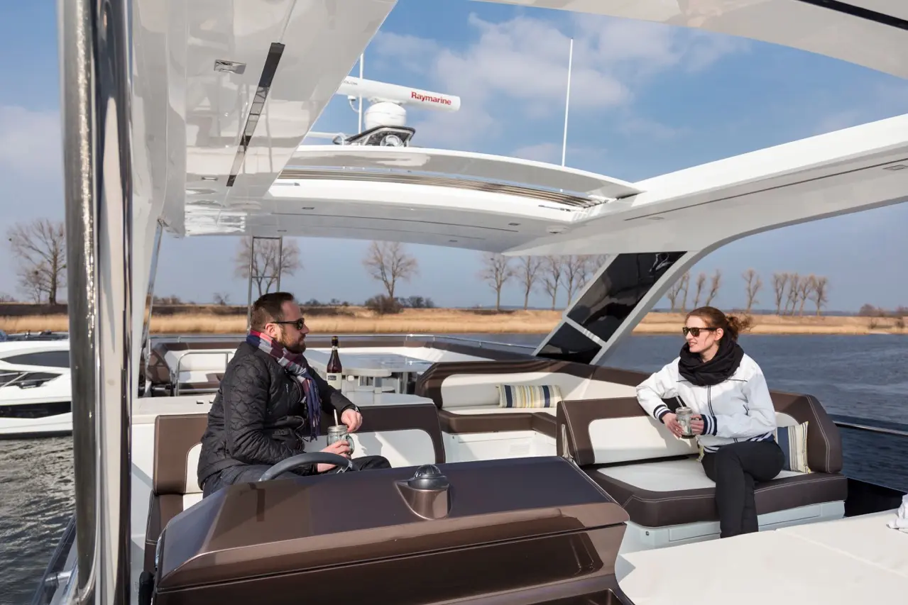 Two people relax on a luxury boat, enjoying drinks while surrounded by water and trees under a clear sky.