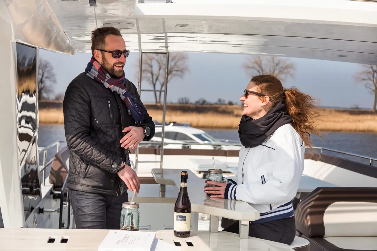 A man and woman enjoy a drink on a yacht, smiling at each other, with scenic river views and a bottle of beer on the table.
