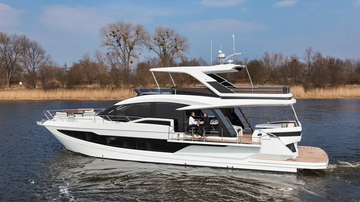 A luxury yacht gliding on water with two people seated on the deck, surrounded by trees and a clear blue sky.