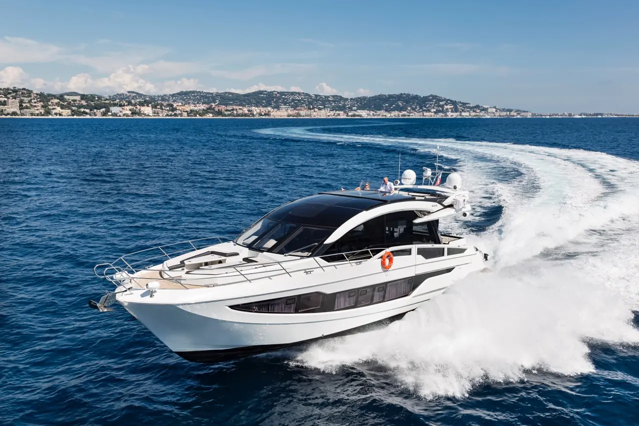 Luxury motor yacht speeding over blue water with coastline in the background, showcasing sleek design and summer leisure.
