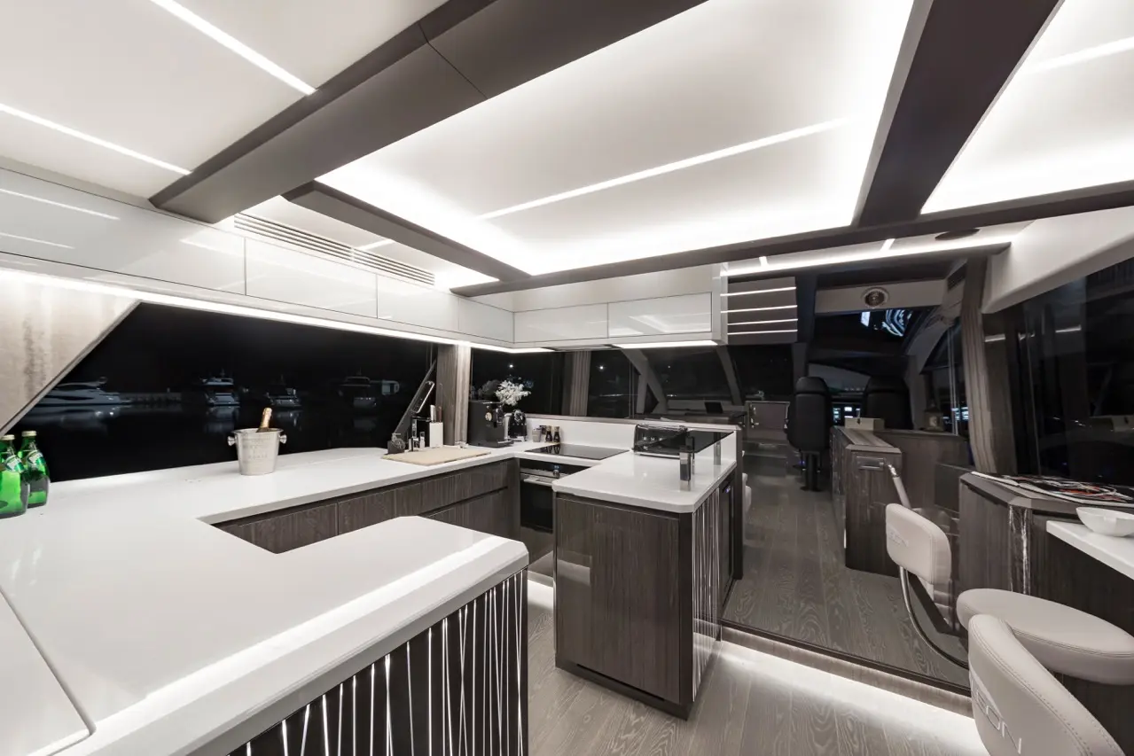 Modern yacht kitchen interior featuring white countertops, dark cabinetry, and stylish lighting, with a view of boats outside.