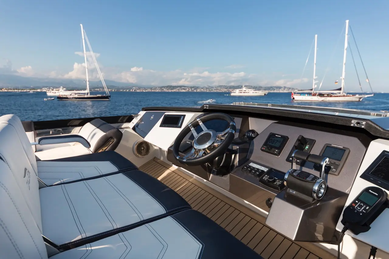 Luxury yacht interior with steering wheel and control panel; sailboats and coastline visible in background under clear sky.