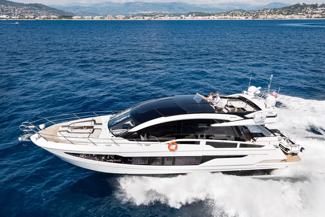 Luxury yacht cruising on blue waters, with two people on deck, mountains and coastline in the background.
