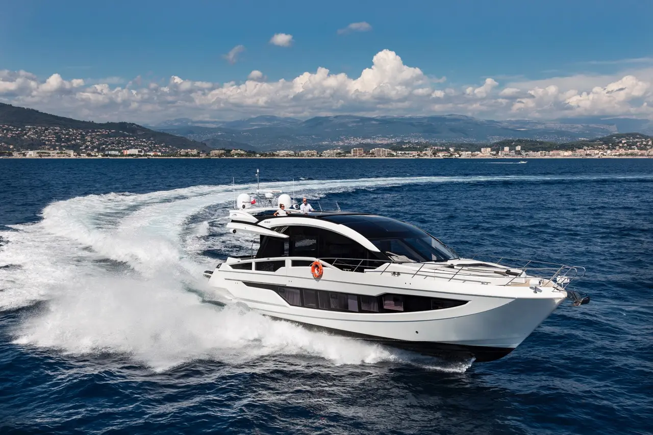 Luxury motor yacht cutting through blue waters, creating white foam, with a coastal backdrop and cloudy sky.