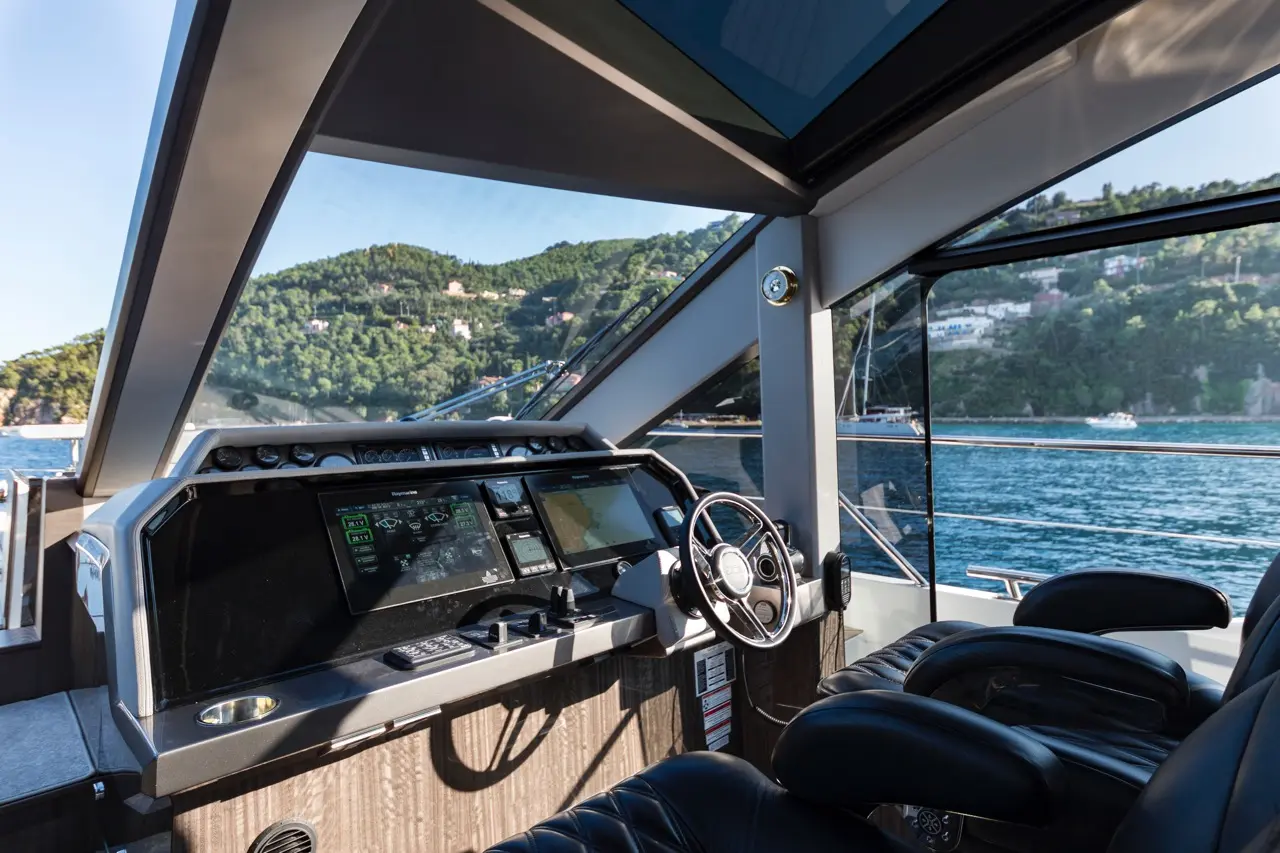 Boat cockpit view with steering wheel, screens displaying navigation data, and scenic coastline in the background.