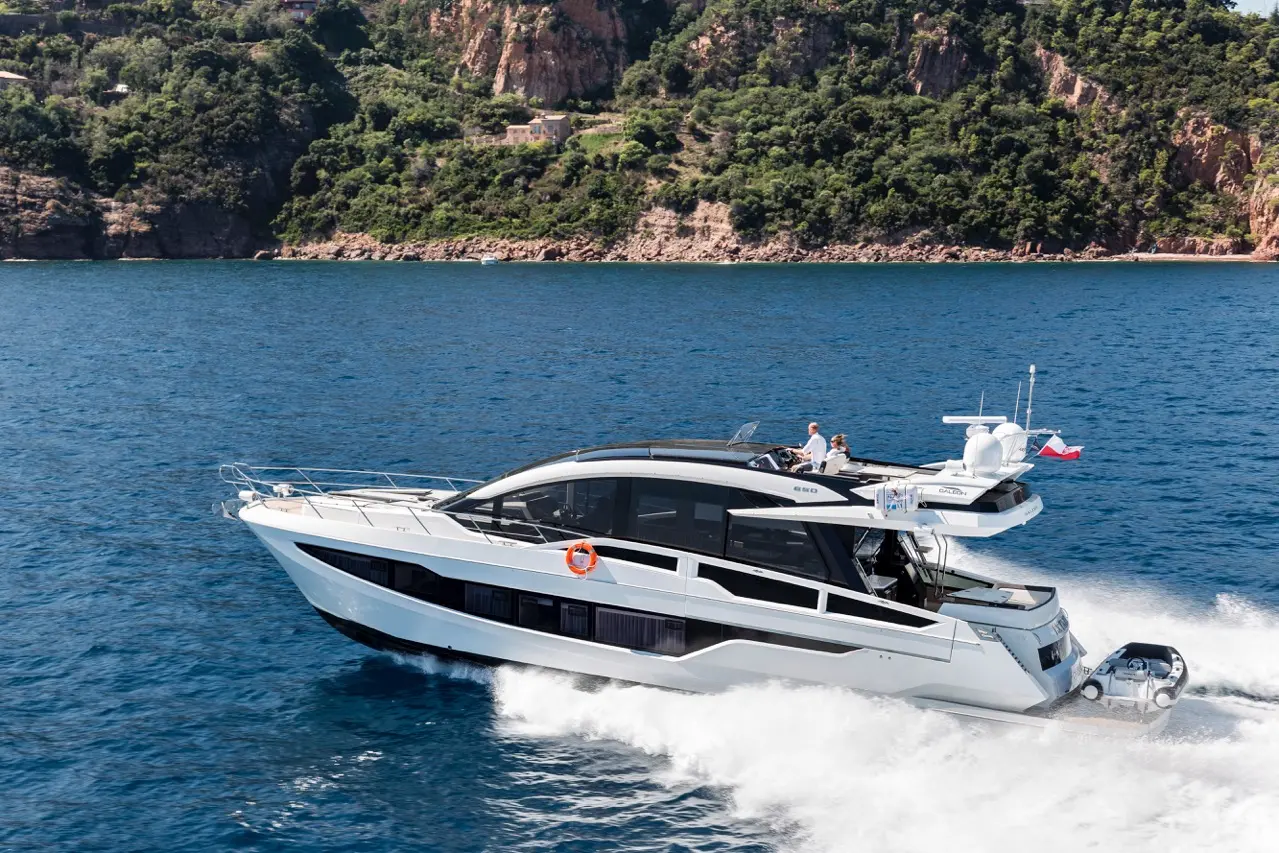 Luxury yacht cruising on blue waters against a lush green hillside, featuring two people on board enjoying the ride.