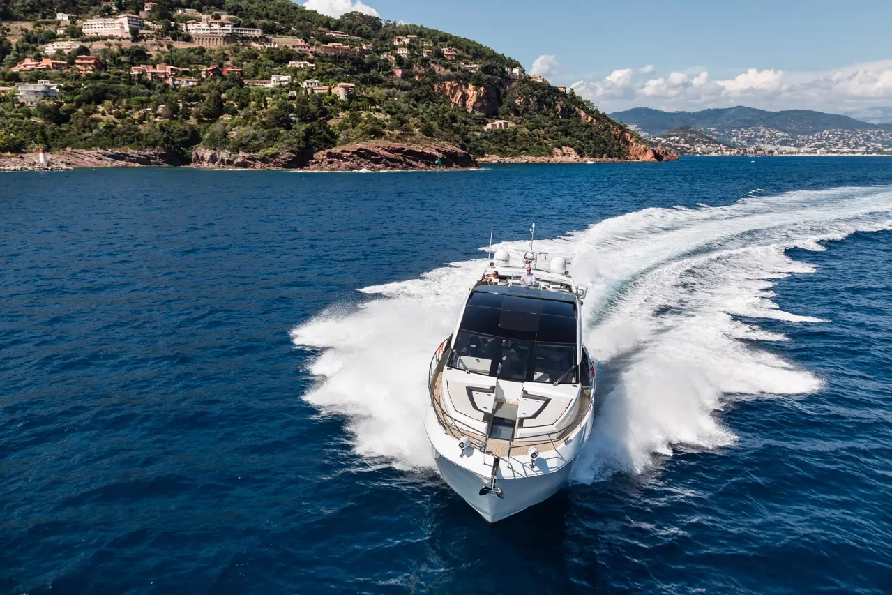 Luxury motorboat cruising on blue waters near a rocky coastline with lush greenery and residential buildings in the background.
