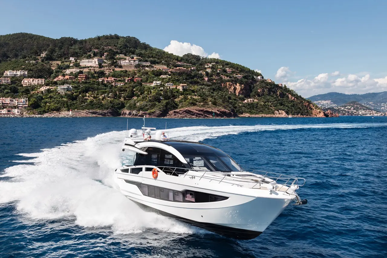 A sleek white motorboat navigates through blue water near a lush green hillside with residential buildings.