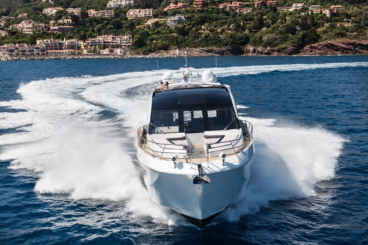 A luxury motorboat cuts through blue waters, leaving a wake, with hillside villas visible in the background.