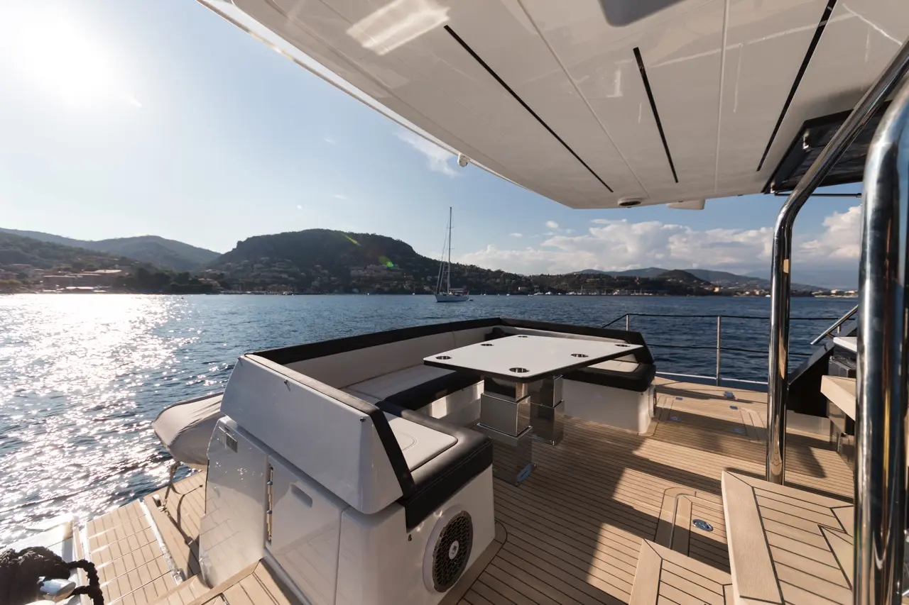 Modern boat deck with a dining area, ocean view, and distant hills under a sunny sky reflecting on the water.