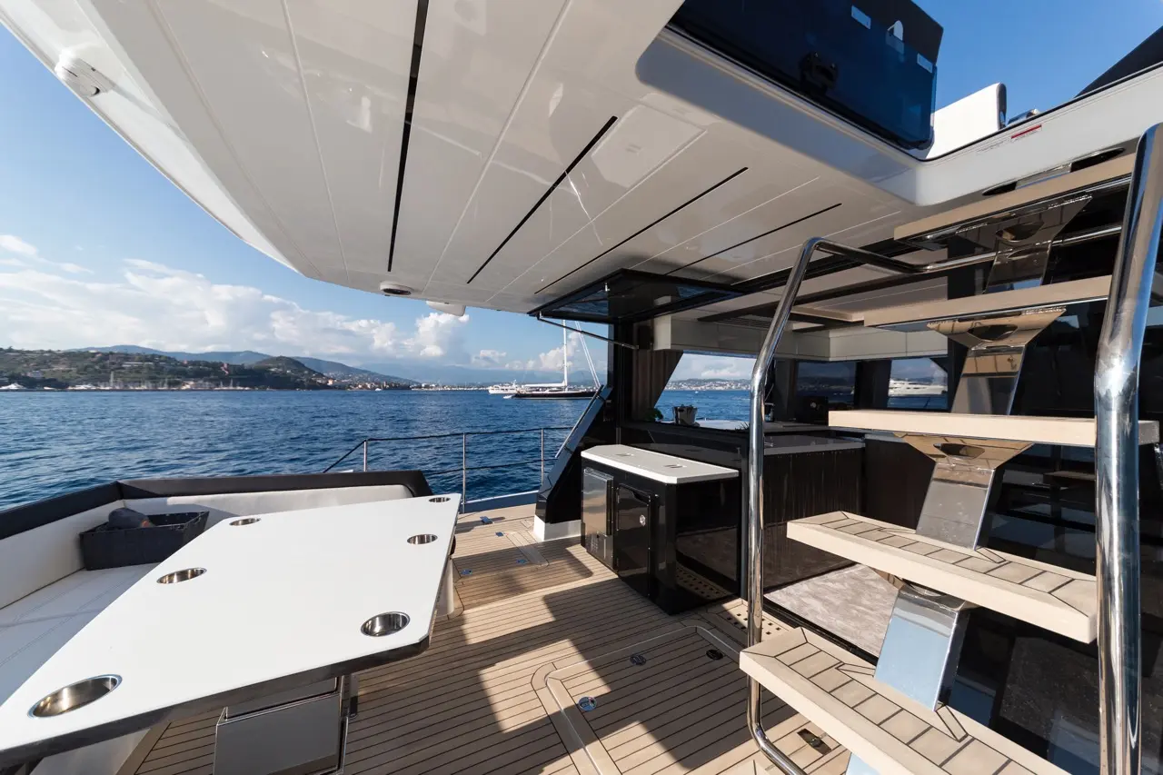 Spacious yacht deck with a dining area, sleek stairs, and a panoramic view of the sea and coastal landscape.