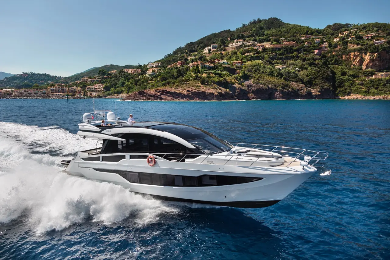 A sleek yacht powered through clear blue waters, with a coastal hillside and luxury homes in the background.